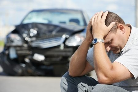 Hombre afligido sentado frente a su auto dañado tras un accidente